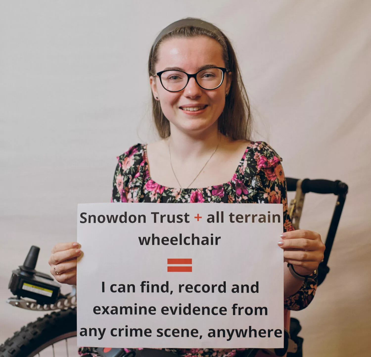 Image of Kira sitting in her powerchair and smiling at the camera. She is holding a sign that says 