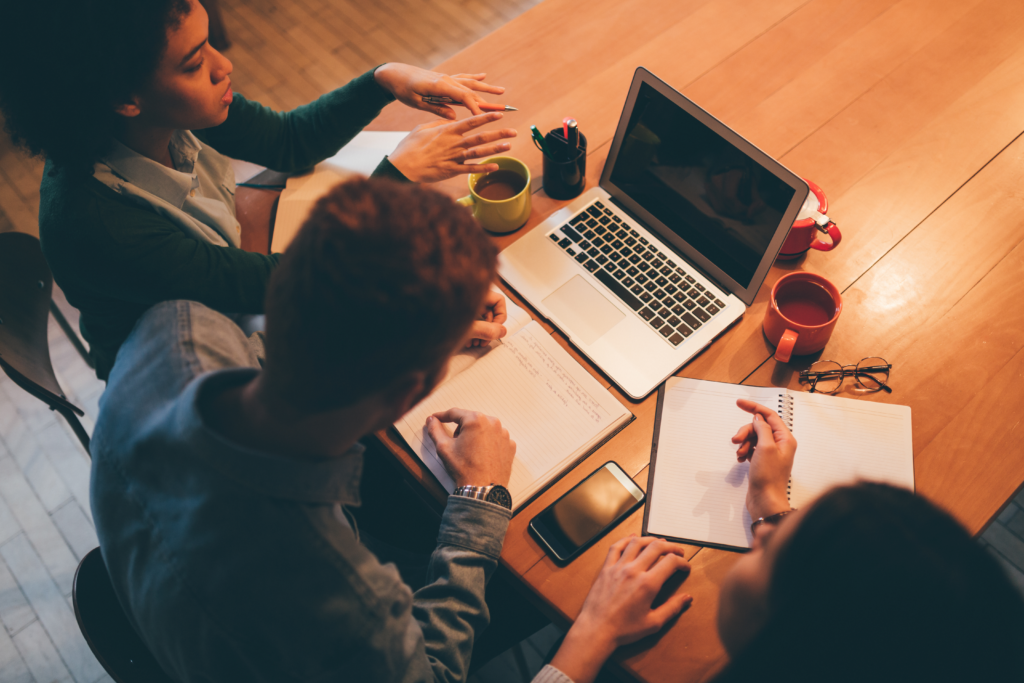 Image of a laptop on a desk surrounded by cups of tea and coffee. Three students are sitting at the desk talking and making notes.