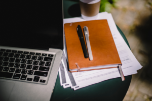 Image of printed notes, a notebook and pens in a pile next to an open laptop