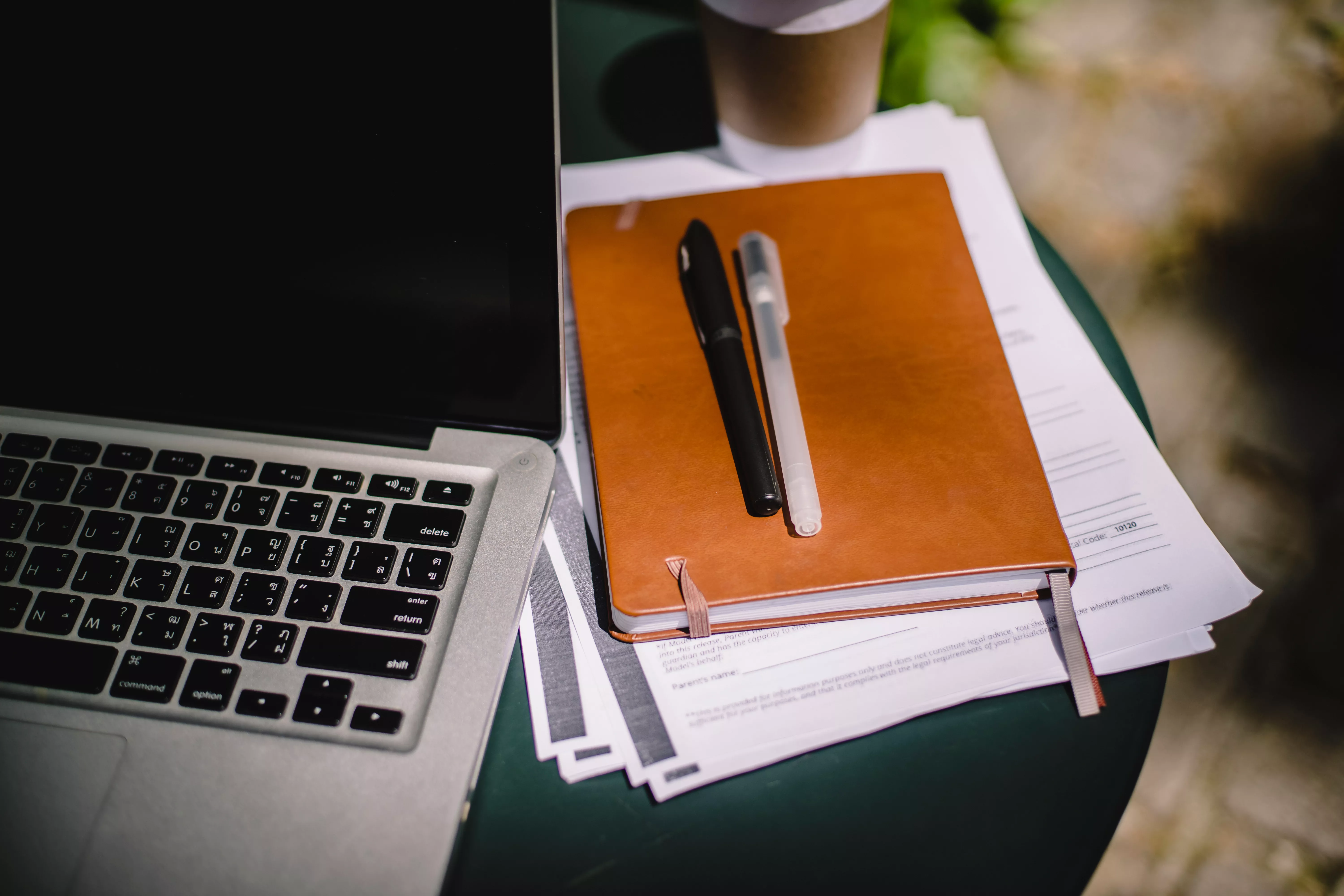 Image of a notebook and two pens on a pile of papers. To the left is an open laptop