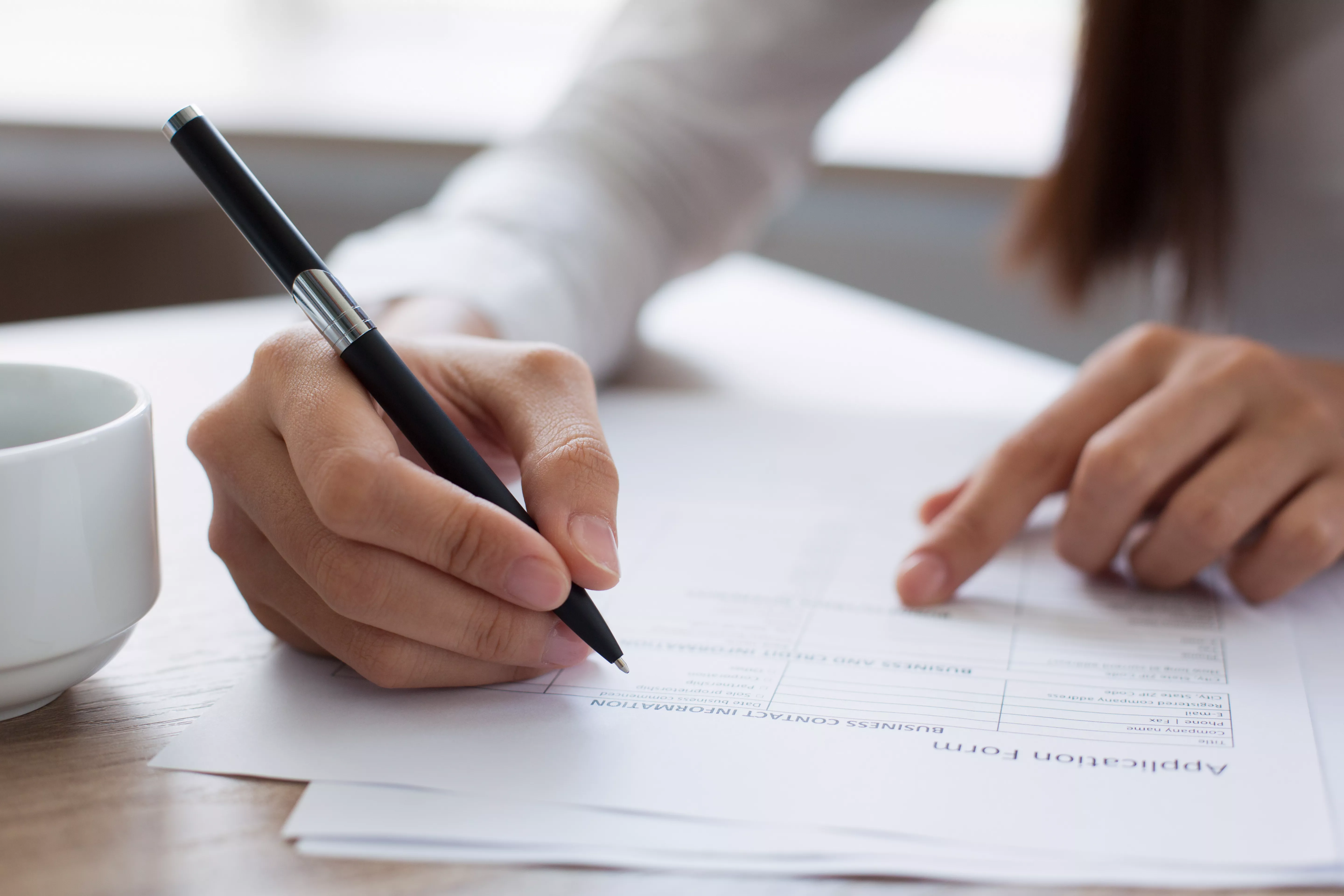 Close-up image of a pair of hands. One is holding a pen and writing on a sheet of paper, while the other is resting on the paper while pointing to something. To the left is a mug of coffee. Image is used to illustrate someone completing an application form