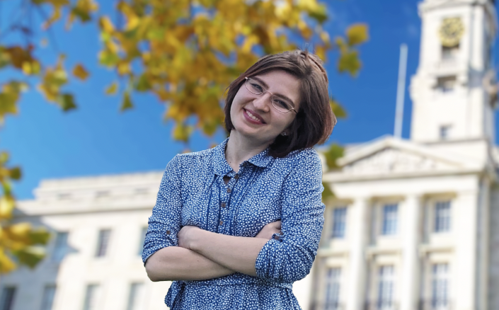 Maria is smiling at the camera with her arms folded. She is wearing a blue dress and glasses, and has shoulder-length brown hair. Maria is a winner of the 2023 Snowdon Master's Scholarships