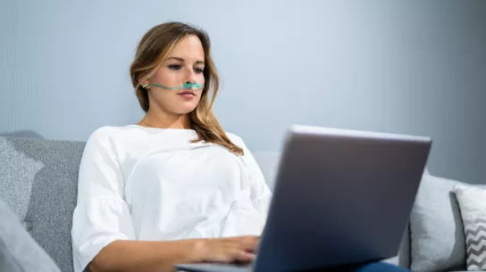 A young woman is sitting on a sofa and using a laptop. She is using a nasogastric tube