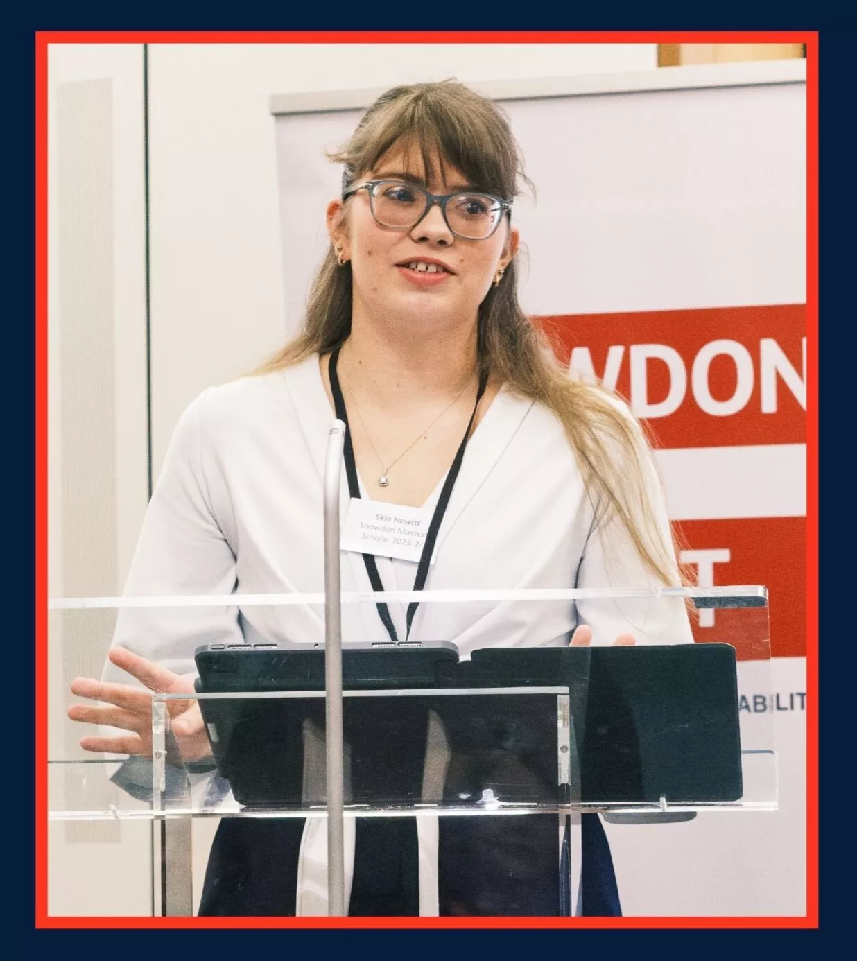 A woman stands at a lectern giving a speech, in front of a background with the Snowdon Trust logo of a red equals sign