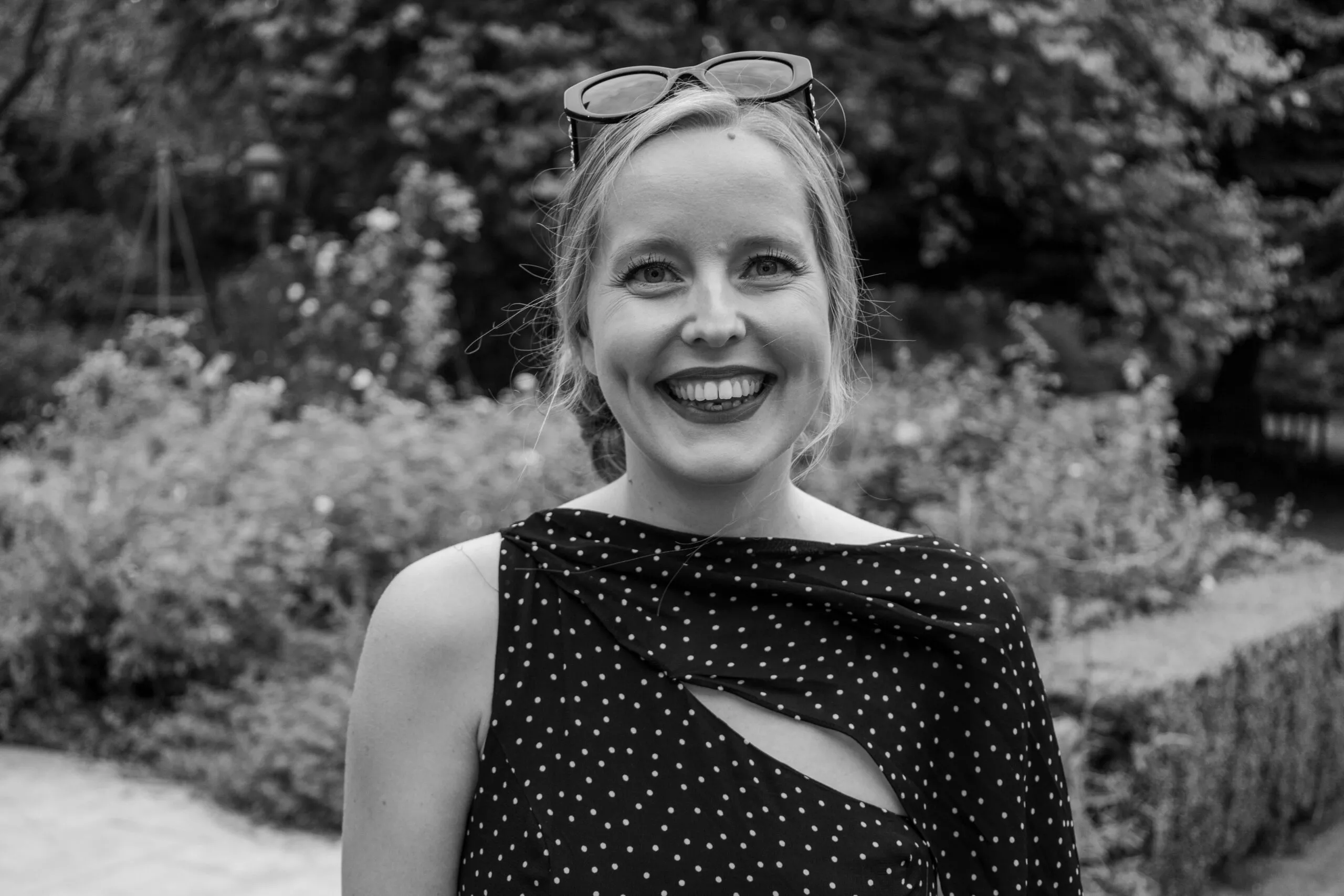 A black and white photo of Amelia who is a white young woman wearing her blonde hair up and smiling into the camera. She is wearing a black and white spotty dress and the photo has been taken at a reception in the grounds of the Ambassador’s Residence in Paris.